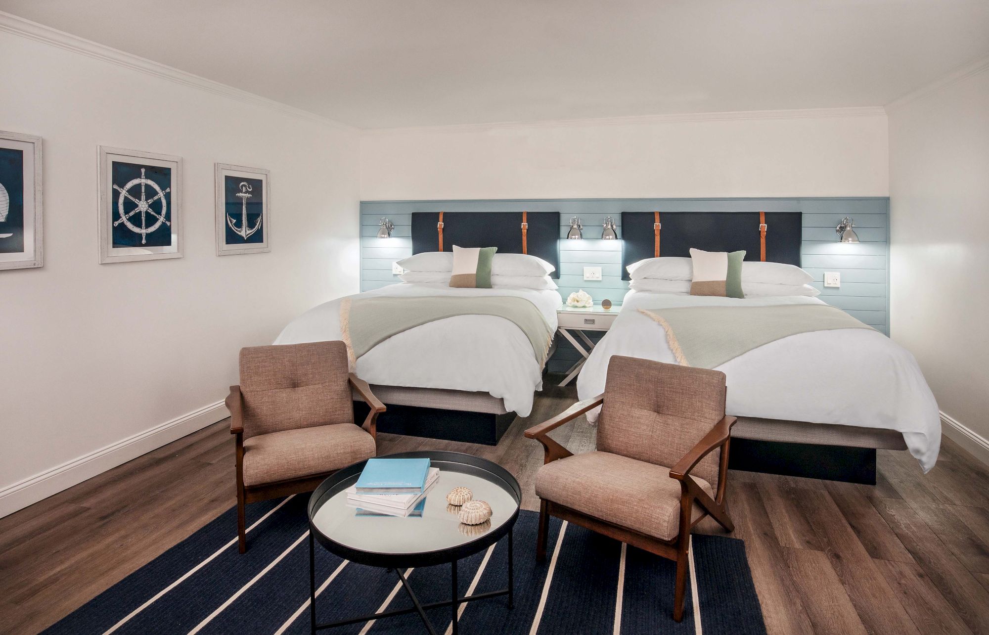 A modern hotel room with two beds, two chairs, a round table, and nautical-themed decor on the walls, featuring a calm and cozy ambiance.