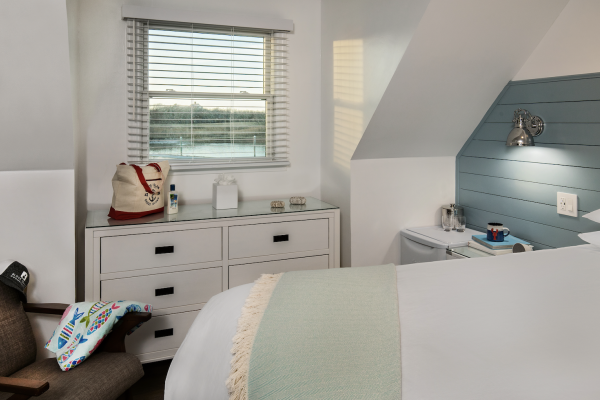 This image shows a neatly arranged bedroom with a bed, a dresser with items on top, a small table with a lamp, and a window with blinds.