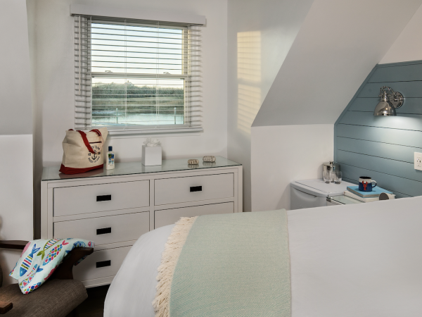 This image shows a neatly arranged bedroom with a bed, a dresser with items on top, a small table with a lamp, and a window with blinds.
