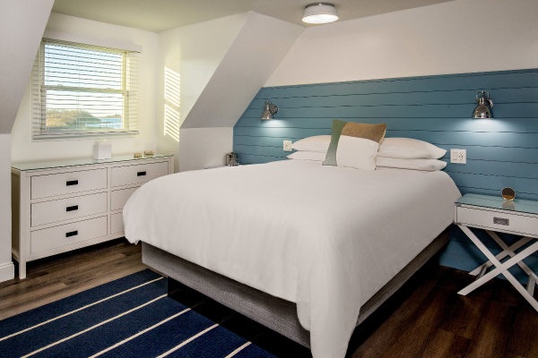 A cozy bedroom with a white bed, blue accent wall, bedside tables, dresser, window with blinds, and a striped rug on a wooden floor ending the sentence.