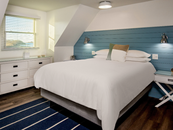 A cozy bedroom with a white bed, blue accent wall, bedside tables, dresser, window with blinds, and a striped rug on a wooden floor ending the sentence.