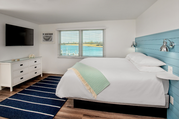 A cozy bedroom with a large bed, dresser, wall-mounted TV, and a window offering a scenic lake view. A blue-striped rug adds a touch of color.