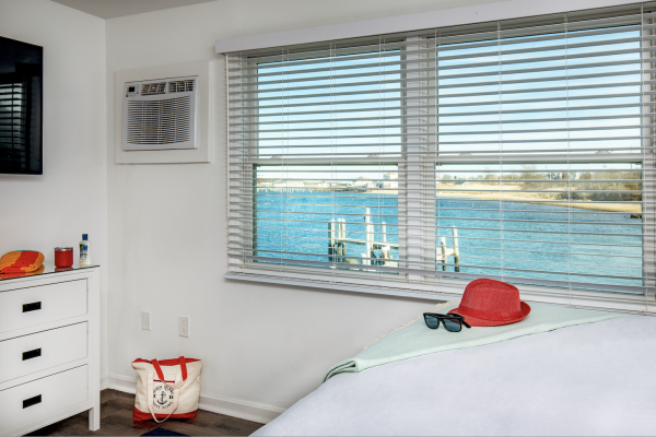 A room with a view of a lake, a dresser with items, a bed with a hat and sunglasses, and a tote bag on the floor next to the wall.