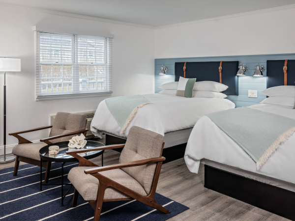 This image shows a neatly arranged bedroom with two beds, two chairs, a small table, a floor lamp, and a window with blinds.