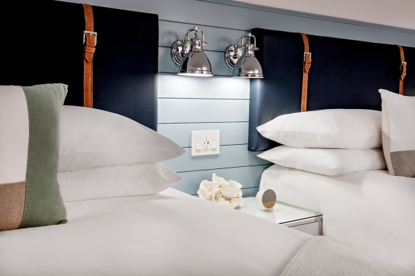 The image shows a hotel room with two neatly made beds, a nightstand with decor, dual wall-mounted lamps, and a shiplap headboard.