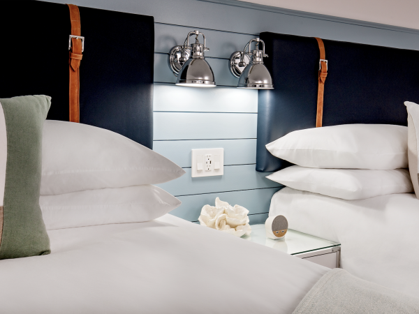 The image shows a hotel room with two neatly made beds, a nightstand with decor, dual wall-mounted lamps, and a shiplap headboard.