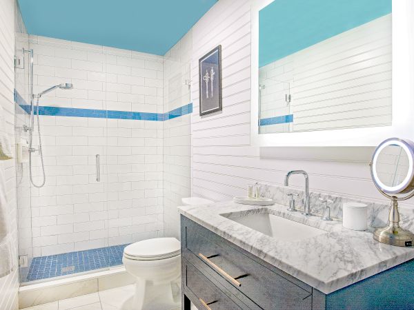 A modern bathroom features a marble countertop, mirror with lights, vanity, towel rack, toilet, shower with glass door, blue tiles, and wall art.