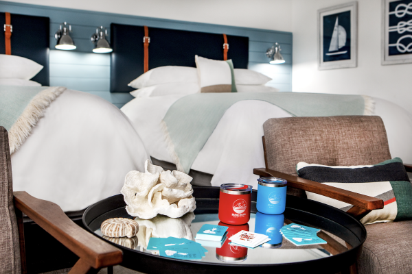 The image shows a hotel room with two beds, a round table with two cans, playing cards, and seashells, and brown chairs.