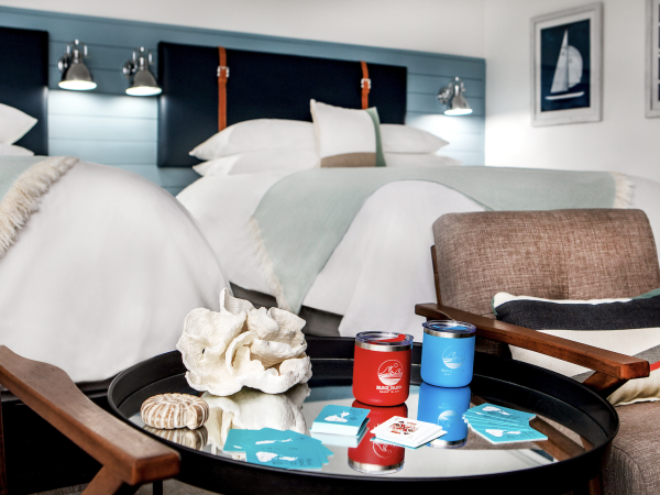 The image shows a hotel room with two beds, a round table with two cans, playing cards, and seashells, and brown chairs.