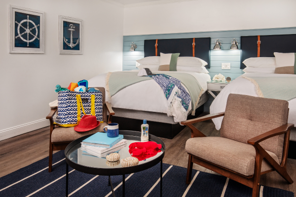 A cozy room with two beds, nautical-themed decor, a chair, and a table with beach items like sunscreen, a hat, and seashells, suggesting a beach getaway.