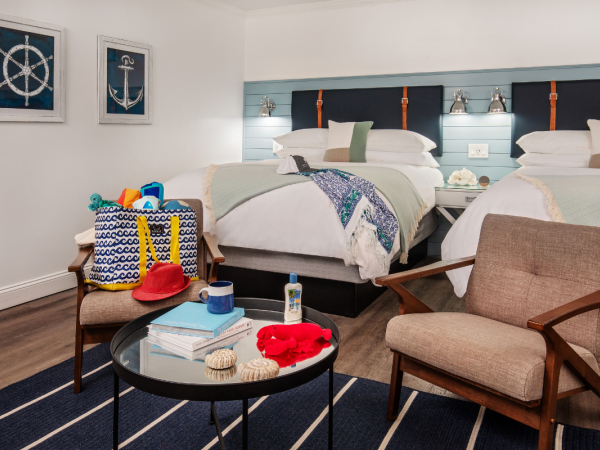 A cozy room with two beds, nautical-themed decor, a chair, and a table with beach items like sunscreen, a hat, and seashells, suggesting a beach getaway.