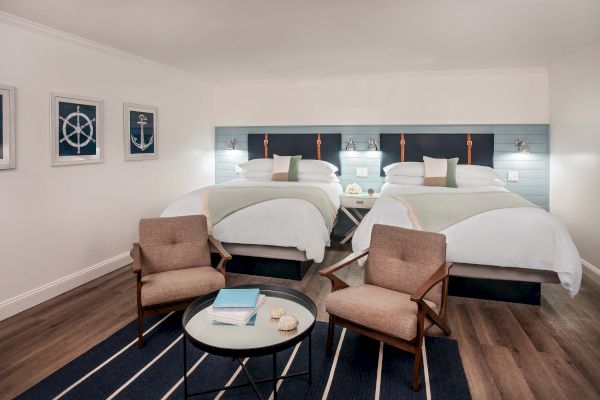 The image shows a hotel room with two double beds, two armchairs, a round table, and nautical-themed wall art on a light wood floor.