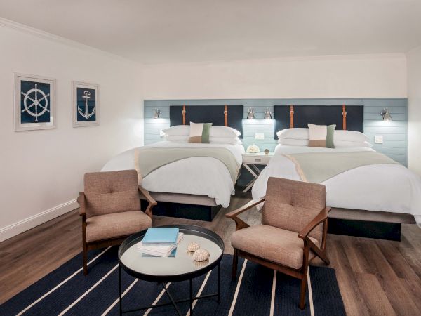 The image shows a hotel room with two double beds, two armchairs, a round table, and nautical-themed wall art on a light wood floor.