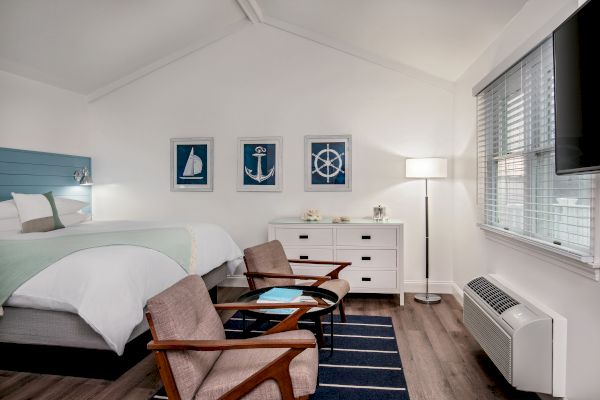 A cozy bedroom with nautical decor, including bed, chairs, dresser, and wall art of a sailboat, anchor, and ship wheel, blue accents throughout.