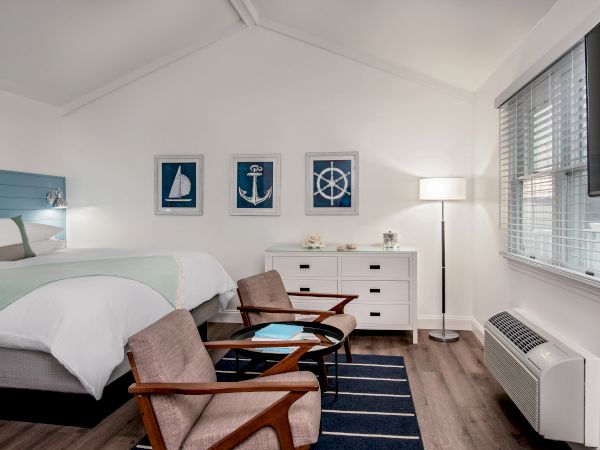 A cozy bedroom with nautical decor, including bed, chairs, dresser, and wall art of a sailboat, anchor, and ship wheel, blue accents throughout.
