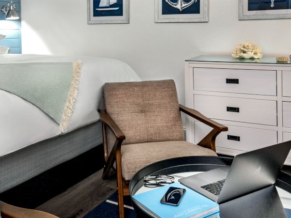 A cozy room features a bed with a light blue blanket, a dresser, a laptop on a round table, and two chairs, decorated with nautical-themed artwork.