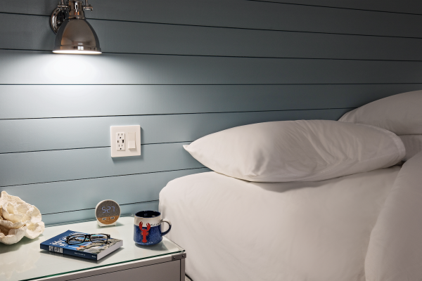 A cozy bedroom setup with a bed, a wall lamp, a smart speaker, a book, a pair of glasses, and a coffee mug on the bedside table.