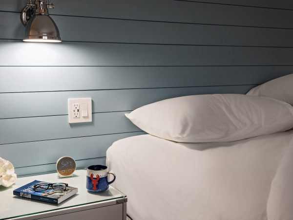 A cozy bedroom setup with a bed, a wall lamp, a smart speaker, a book, a pair of glasses, and a coffee mug on the bedside table.