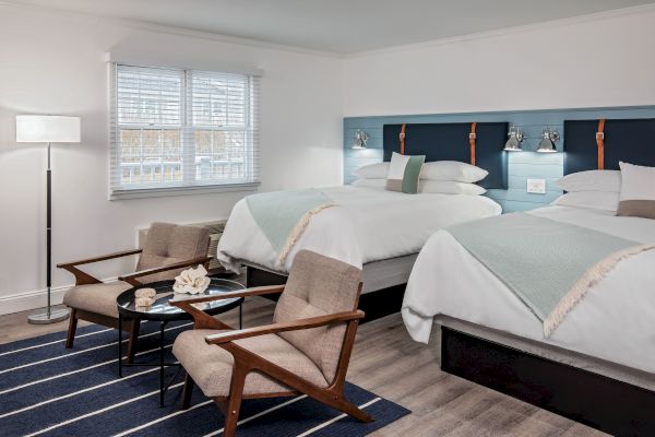 The image shows a clean, modern hotel room with two beds, two chairs, a small table, a floor lamp, and a window with blinds letting in natural light.