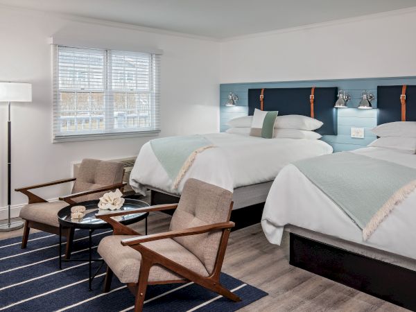 The image shows a clean, modern hotel room with two beds, two chairs, a small table, a floor lamp, and a window with blinds letting in natural light.