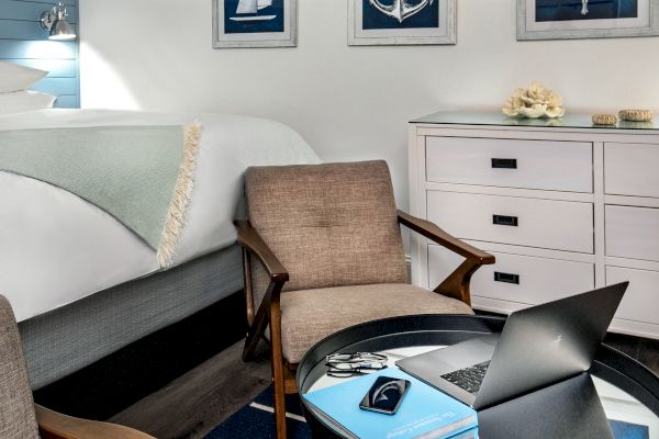 A cozy room with a bed, two chairs, a dresser, and a small table. A laptop, notebook, and phone are on the table. Nautical art on the walls.