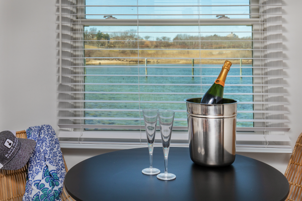 A table by a window with a water view holds a champagne bottle in an ice bucket, two glasses, a hat, and a blue patterned cloth.