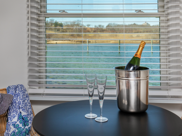 A table by a window with a water view holds a champagne bottle in an ice bucket, two glasses, a hat, and a blue patterned cloth.