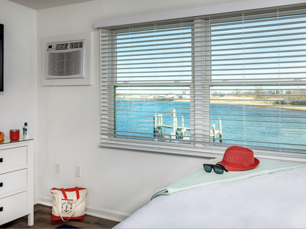 A cozy room with a window overlooking a waterfront, featuring a hat and sunglasses on the bed, and a tote bag on the floor.