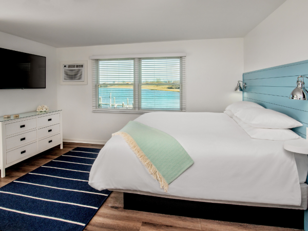 A cozy bedroom featuring a large bed with a green throw, a dresser, a wall-mounted TV, an air conditioner, and a window with a waterfront view.