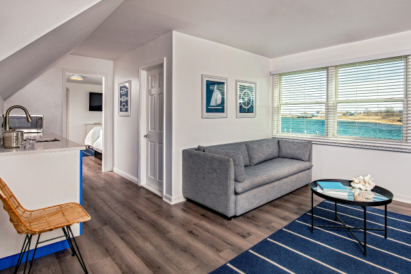 A modern living space features a grey couch, a small round table, wall art, large windows with a view, and a small kitchen area with a barstool.