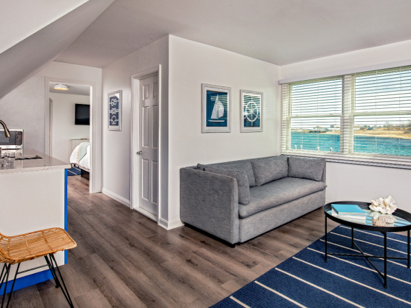 A modern living space features a grey couch, a small round table, wall art, large windows with a view, and a small kitchen area with a barstool.