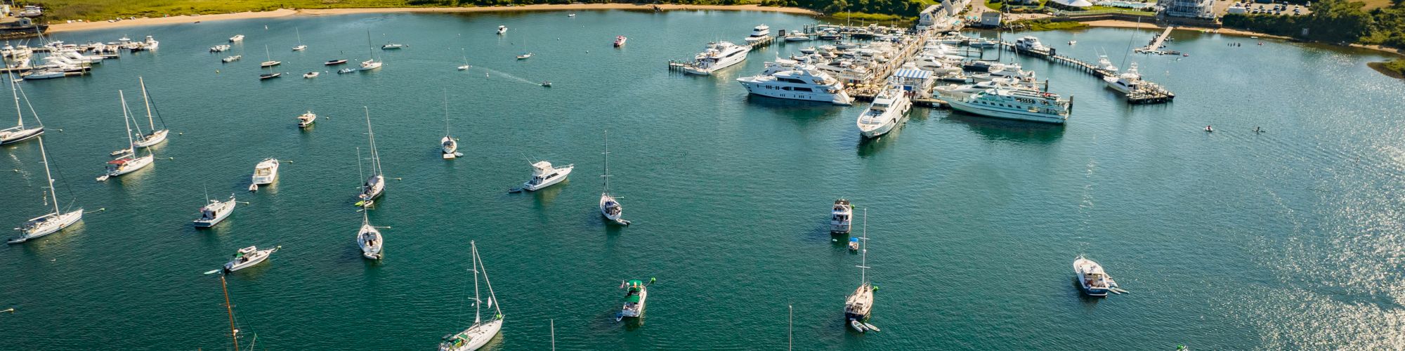 An aerial view of a harbor filled with sailboats and yachts, with a small town and green landscape in the background.