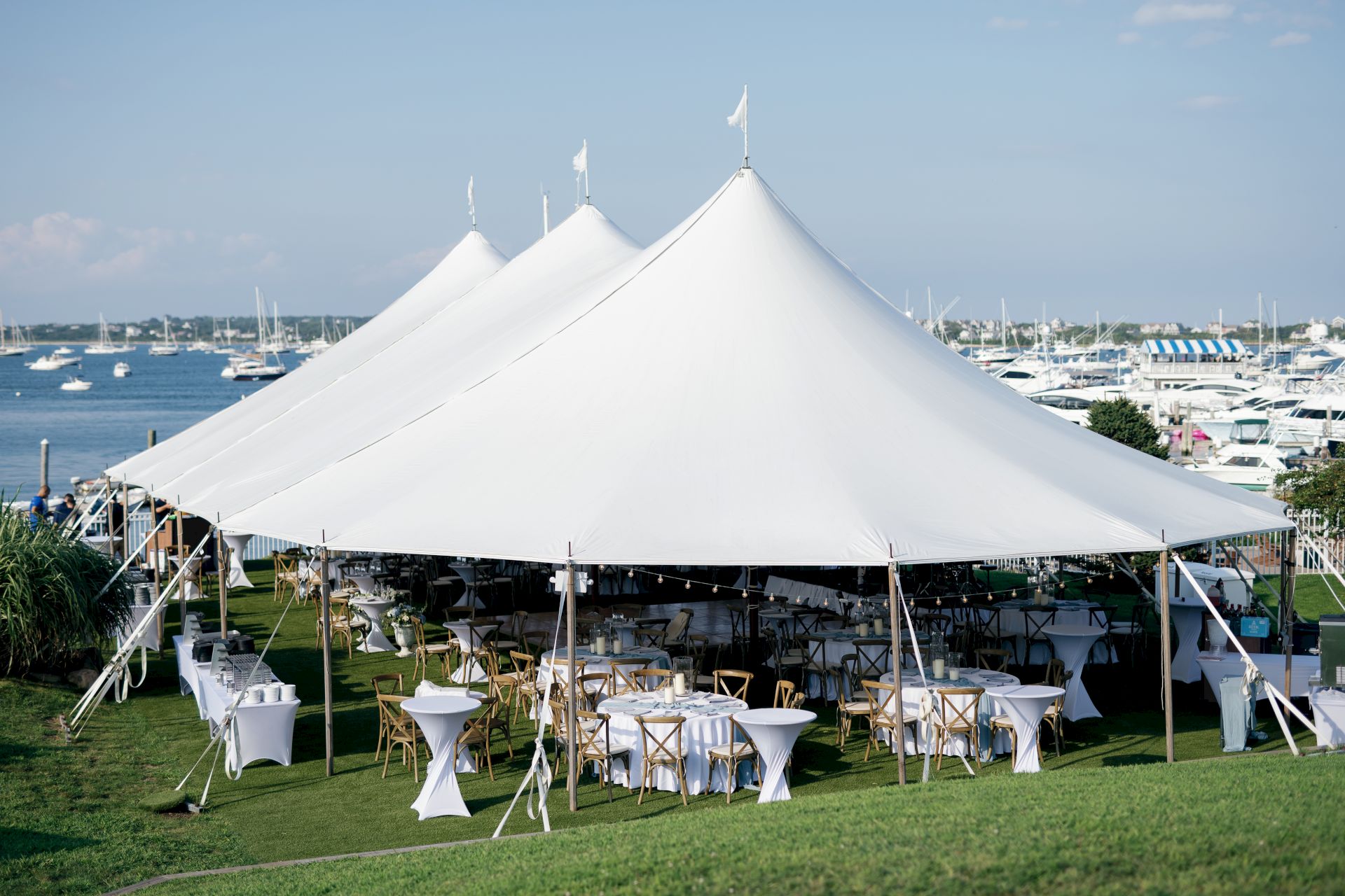 A large white tent is set up on a grassy area by the water, with tables and chairs underneath, likely for an outdoor event.