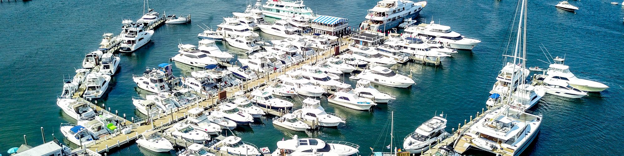 Boats and yachts docked at a large marina, surrounded by a vast expanse of water with more boats anchored in the distance.