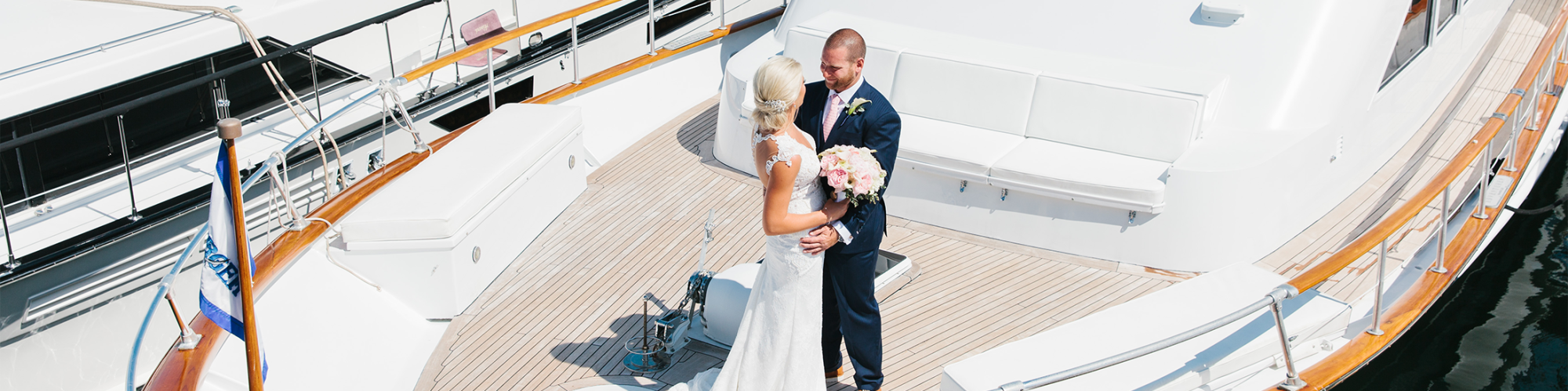 A bride and groom stand on the deck of a boat holding a bouquet, with another boat docked beside them, on a sunny day.