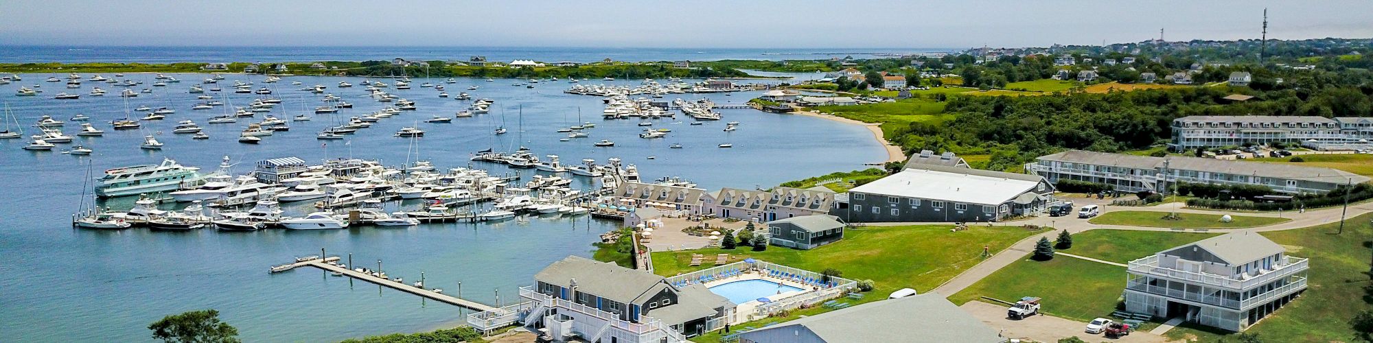 An aerial view of a coastal marina with boats docked, buildings, and a pool surrounded by green landscape under a clear sky.