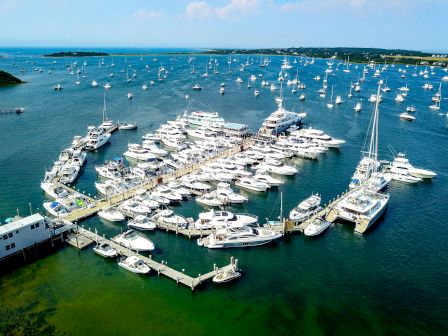 The image shows a marina with numerous boats and yachts docked, surrounded by water and extending piers, under a clear sky.