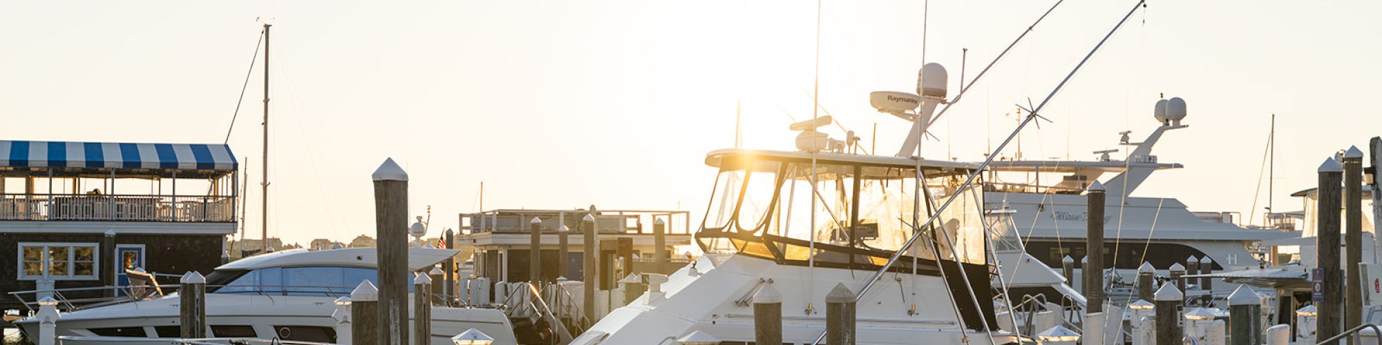 A marina with several boats docked at piers during sunset, with one boat prominently in the foreground and an American flag visible.