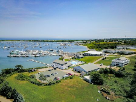 Aerial view of a marina with numerous boats docked, surrounded by green landscape, buildings, and a large parking area. Blue sky overhead ends the sentence.