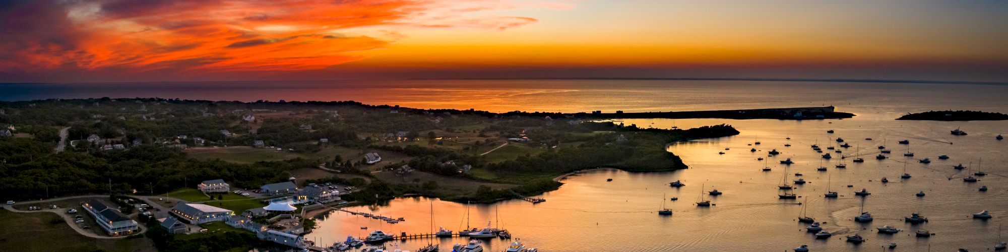 Aerial view of a marina at sunset, with numerous boats docked and anchored, and a dramatic sky with vibrant colors reflecting on the water.