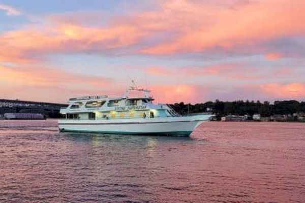 A boat cruises on calm waters during a vibrant sunset, with a bridge and shoreline in the background.