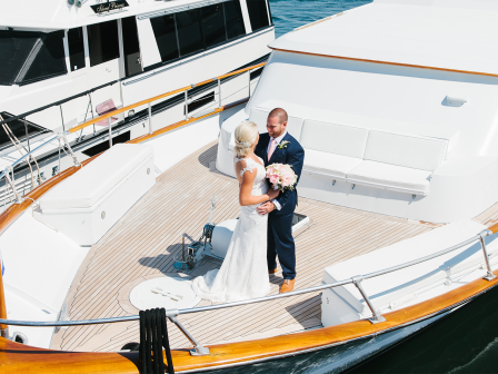 A couple in wedding attire stands on the deck of a yacht, embracing and holding a bouquet, with another boat docked nearby.