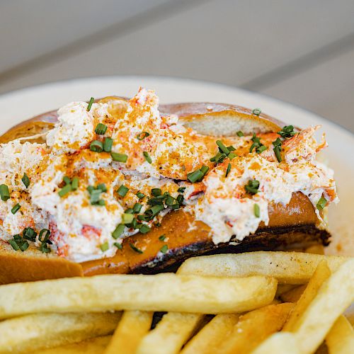 The image shows a lobster roll topped with a creamy mixture, garnished with green herbs, and served with a side of French fries on a white plate.