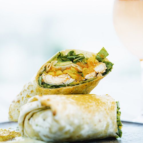 The image shows two chicken wraps filled with vegetables, placed on a plate, with a glass of a light-colored beverage in the background.