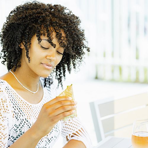 A person with curly hair is looking at a wrapped snack while sitting outdoors beside a glass of juice on a table, wearing a white crocheted top.