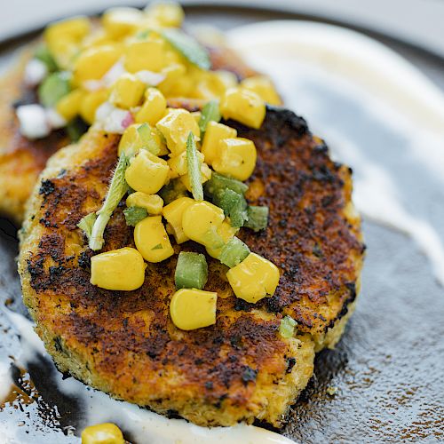 Two golden-brown patties topped with a corn and vegetable mix, served on a plate with a swirled white sauce garnish.