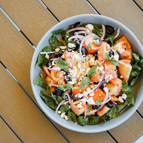 The image shows a fresh salad with lettuce, tomatoes, feta cheese, red onions, black olives, and herbs in a bowl on a wooden surface.