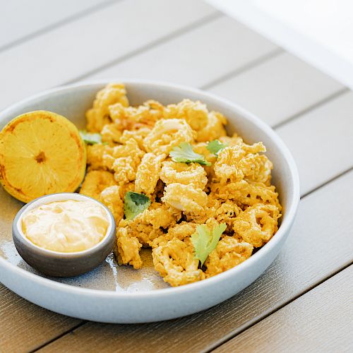 A plate of fried calamari garnished with herbs, a dipping sauce, and a grilled lemon slice, placed on a wooden table.