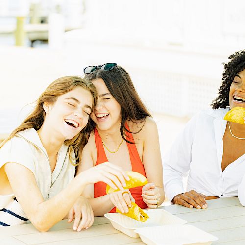 Three women sitting at a table outdoors, laughing and eating tacos.