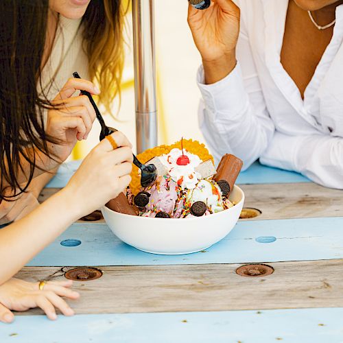 Three people are enjoying a large bowl of ice cream topped with various toppings while sitting around a blue and wooden picnic table.